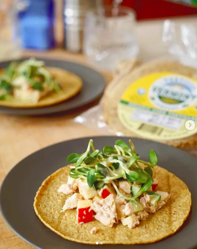 Open corn tortilla topped with tuna salad, diced red apple and sunflower sprouts on a dark plate