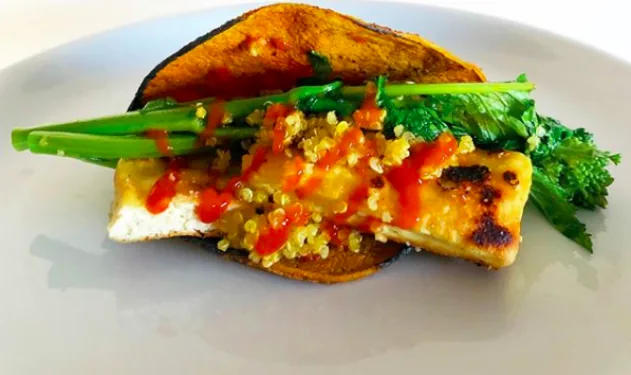 A turmeric corn tortilla holding golden tofu, quinoa and green vegetables with a drizzle of hot sauce