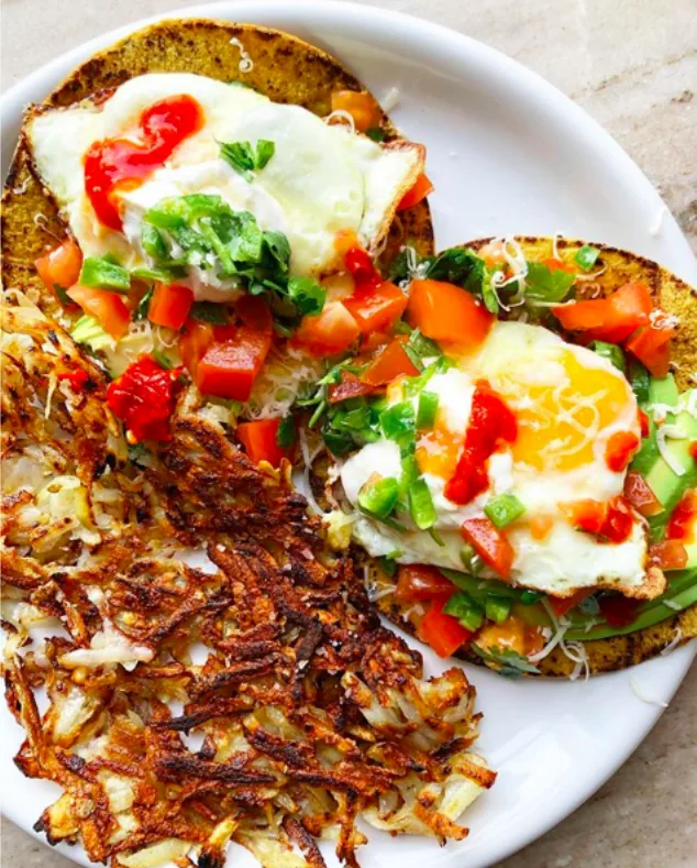 Two corn tortillas topped with fried eggs, avocado and chopped vegetables beside golden hash browns