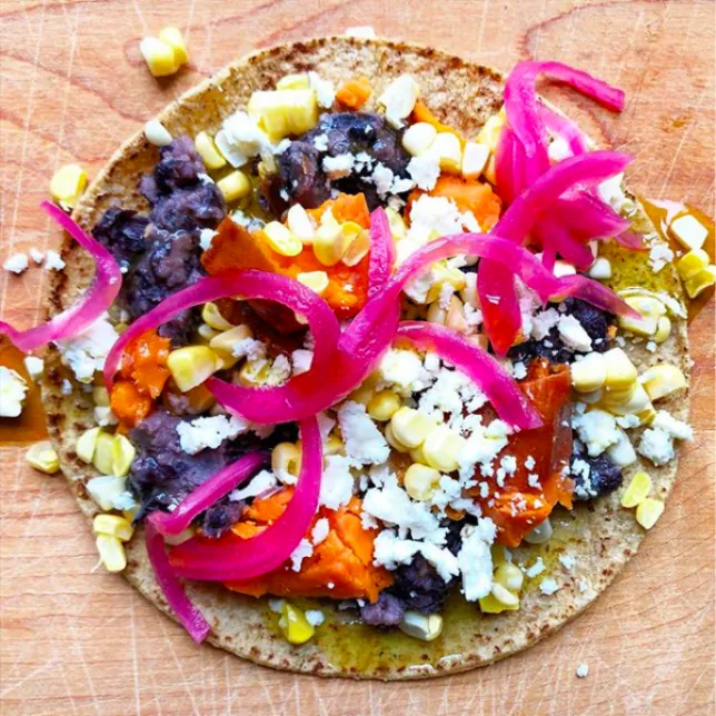 A corn tortilla topped with black beans, orange sweet potato, corn kernels, crumbled queso fresco and pink pickled onions
