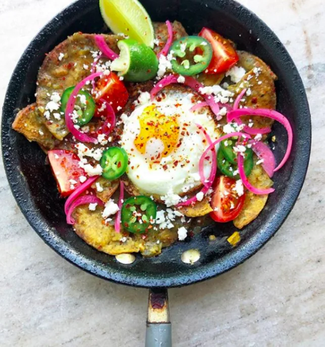 Skillet of salsa-coated corn tortilla pieces with a fried egg, crumbled cheese, jalapeño, tomato, pickled onion and lime