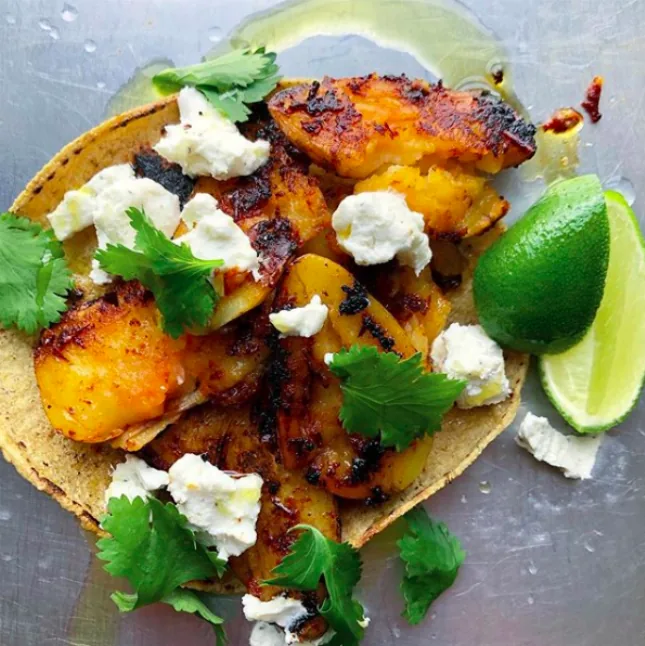 A corn tortilla filled with crisp golden smashed potatoes, crumbled white cheese and cilantro beside lime wedges