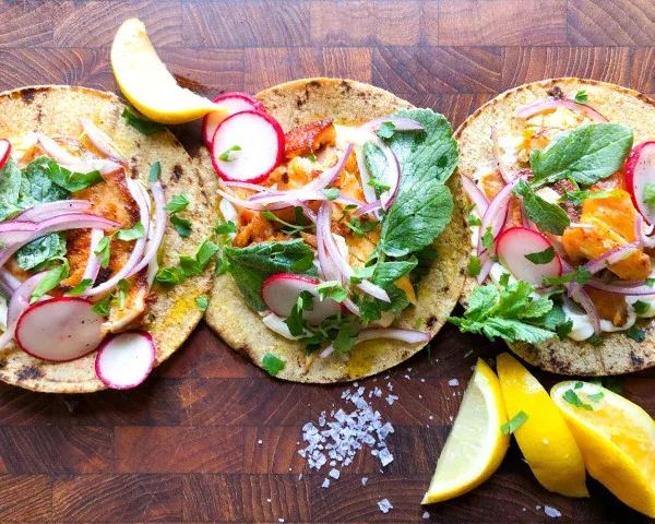 Three corn tortilla tacos topped with vegetables, sliced radishes, onion and fresh herbs