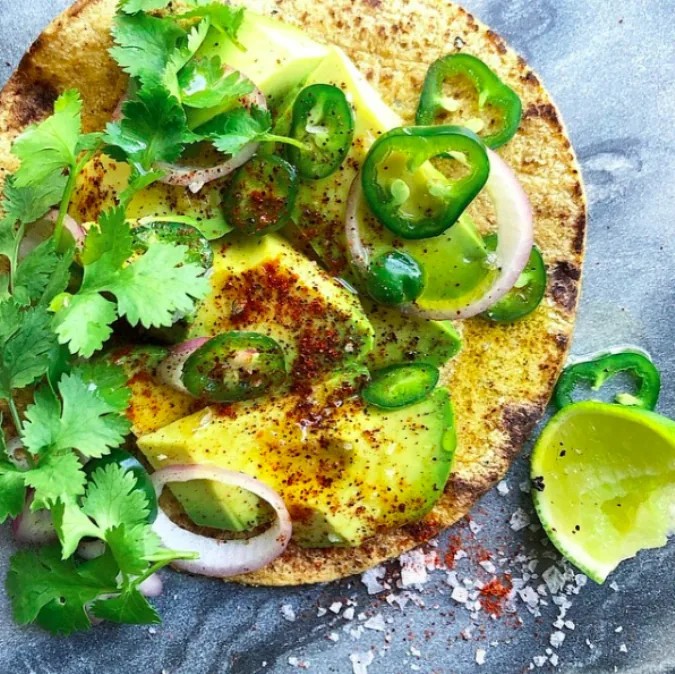 Corn tortilla topped with avocado slices, shallot rings, jalapeño, chili powder and cilantro beside a lime half
