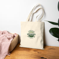 Classic Emblem natural canvas tote, resting on a wooden bench beside a basket and plant